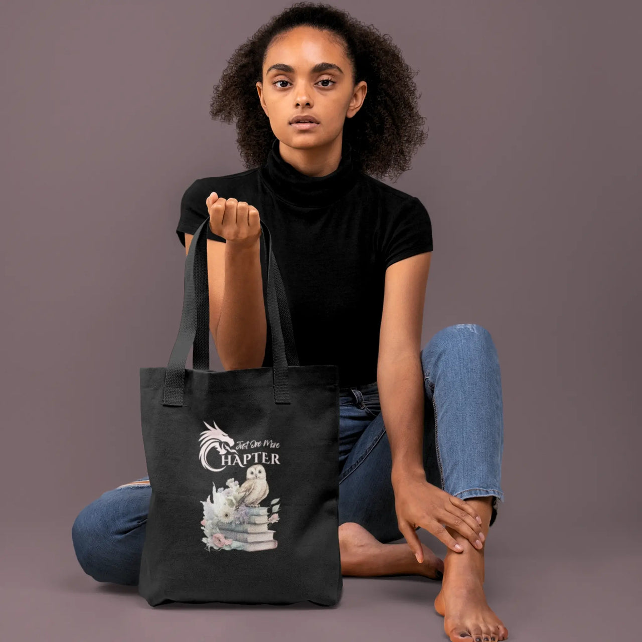 a woman sitting with a Just One More Chapter Owl and Book design on a black tote