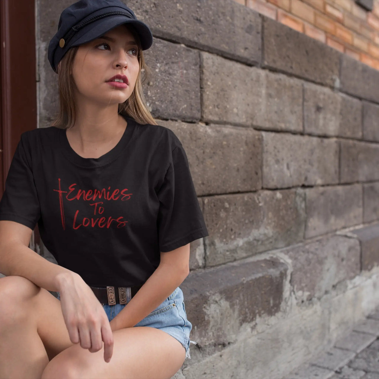 woman crouching next to a wall in a black enemies to lovers tee with red text