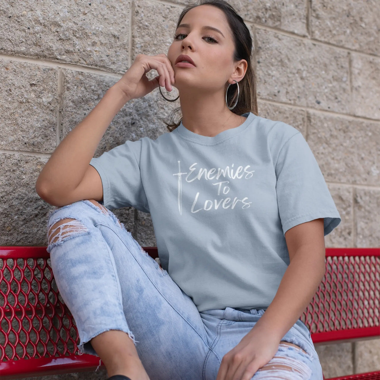 woman sitting on a bench ina light blue Enemies To Lovers Tshirt with white text