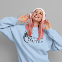 Thumbnail for smiling woman wearing a light blue Just One More Chapter sweatshirt with black text