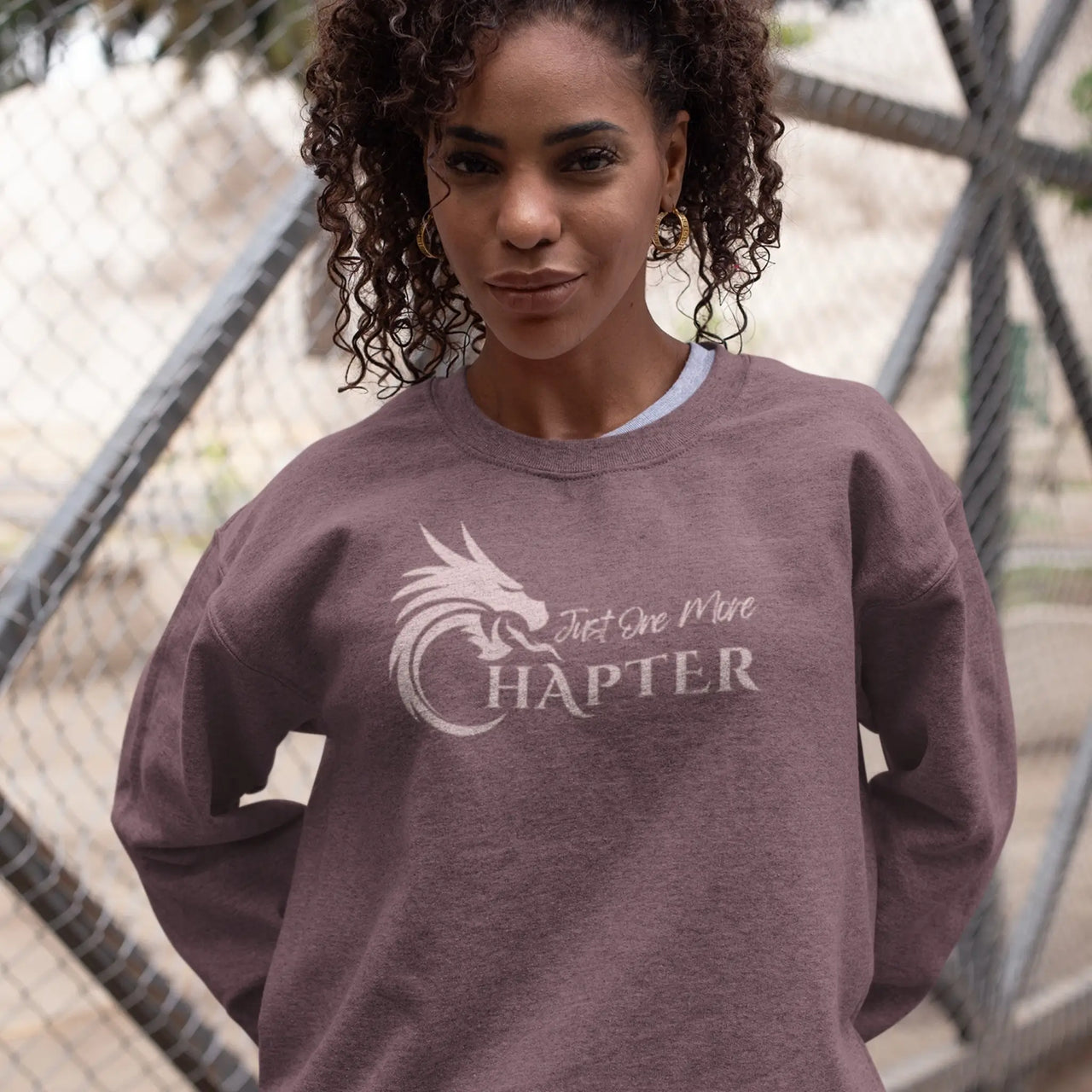 woman wearing a heather maroon Just One More Chapter sweatshirt with pink text