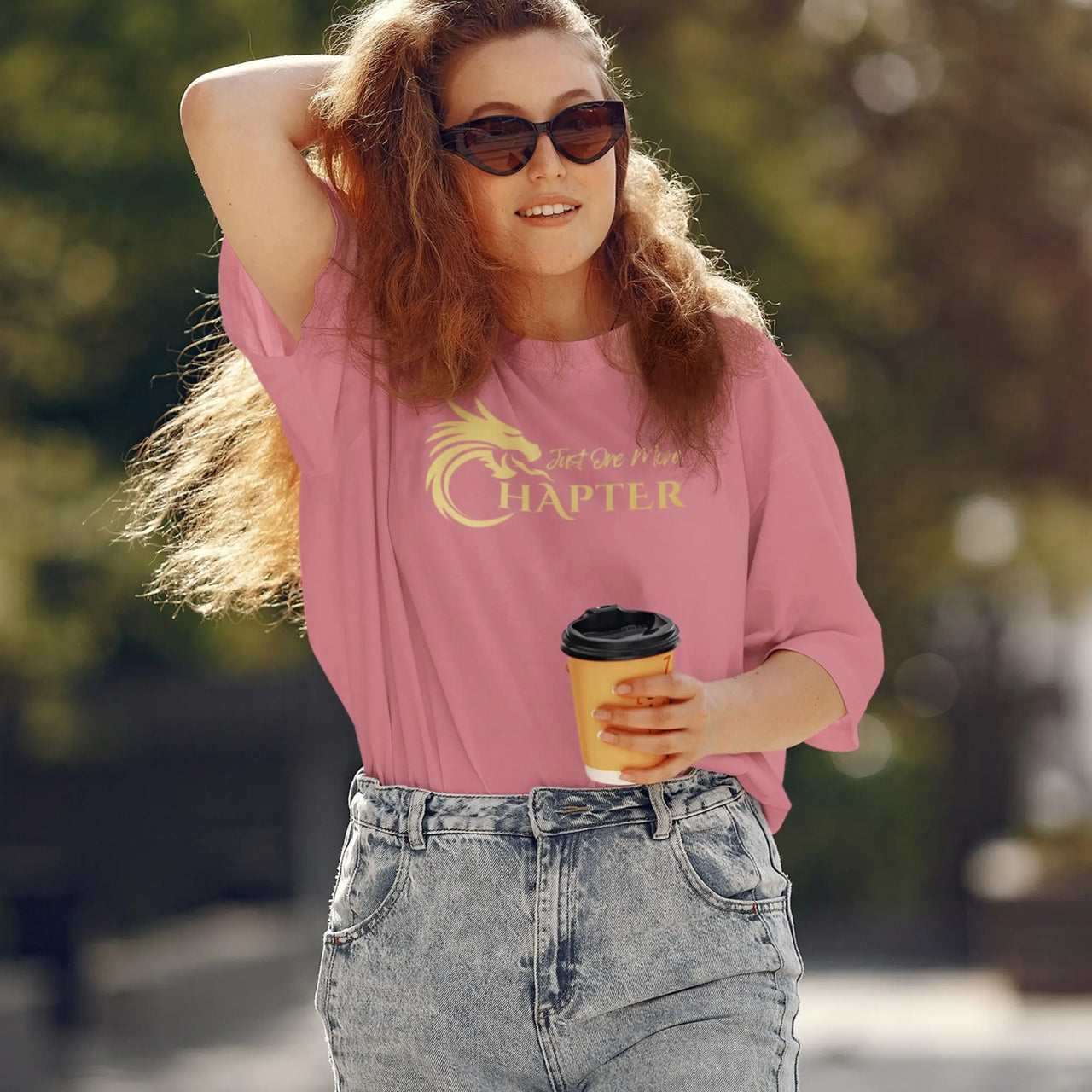 woman drinking coffee weaing a dark pink Just one more chapter tshirt with gold text